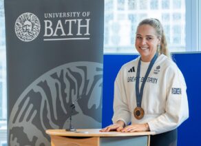 Rower Becky Wilde, wearing an Olympic bronze medal around her neck, standing behind a rostrum and next to a University of Bath banner