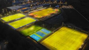 An aerial shot of the floodlit hockey pitches, 400m athletics track, infield, 3G pitches, muliti-use games area (MUGA) and Sports Training Village with the University campus and city of Bath on the horizon. 