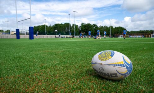 A ground-level photo of the newly-opened 3G pitch at the University of Bath, the first fully recyclable 3G pitch in the UK, with a University of Bath rugby ball in the foreground and players out of focus in the distance