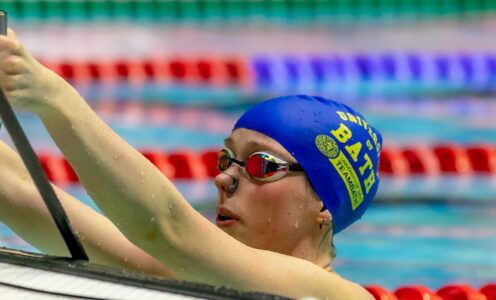 A photo of Niamh Ward, wearing goggles and a blue swimming cap with the words "University of Bath" in gold above the Team Bath logo, holding the rails as she prepares to start a backstroke race at the 2025 BUCS Nationals.