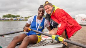 Bath University Boat Club rower Toluwani Adelaja sitting in a single scull boat on the water, next to the pontoon. She is wearing a bronze medal she won at the 2025 British Rowing Club Championships. Crouching down next to her, on the pontoon, is Poppy Kearney, British Rowing Satellite Coach for the South West.