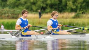 Bath University Boat Club's Archie Norton and Jack Norton rowing on the water in the Open Championship Double Sculls race at the 2025 British Rowing Club Championships. They are wearing blue kit and there are some grass fields in the background. 