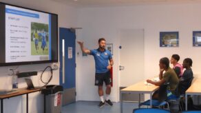 Three footballers, seated, watch as University of Bath Head of Football Che Wilson points to a presentation graphic being displayed on a big screen in a conference room.