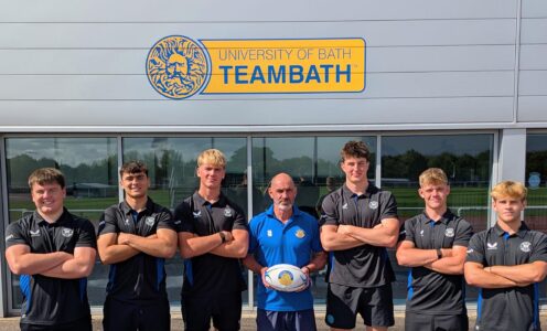 Six Bath Rugby Scholars stand, with their arms crossed, either side of University of Bath Head of Rugby Aaron James, who is holding a rugby ball. They are standing below a Team Bath logo, painted onto the Sports Training Village wall. The players are (from left) Evan Gallagher, Cameron Ward, Fred Kempton, Rory McKnight, Harry Walker and Isaac Mears.