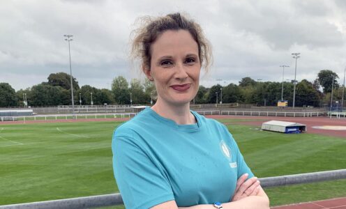 Team Bath senior physiotherapist Katy Williams, wearing a cyan blue Women's Rugby World Cup 2025 t-shirt. She has her arms crossed. Behind her is the outdoor athletics track and field at the University of Bath Sports Training Village.