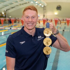 Team GB swimmer Tom Dean holding his three Olympic gold medals in his left hand as he smiles at the camera. Behind him is the London 2012 Legacy Swimming Pool at the University of Bath, where he trained while winning the medals at Tokyo 2020 and Paris 2024.