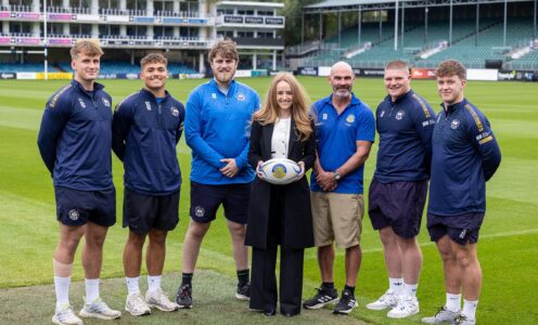 A group of rugby players, coaches and sponsors stand on the pitch at The Rec rugby stadium in Bath. The woman in the middle, Colour Studios Director Rowan Freeman, is holding a rugby ball with the University of Bath logo on it.