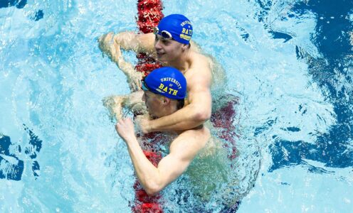Swimmers Matt Ward and Cam Brooker in a pool, resting against the guide rope after a race. Matt, at the top of the picture, has his arm around Cam's shoulder and they are both wearing blue swim caps featuring the University of Bath in gold lettering.