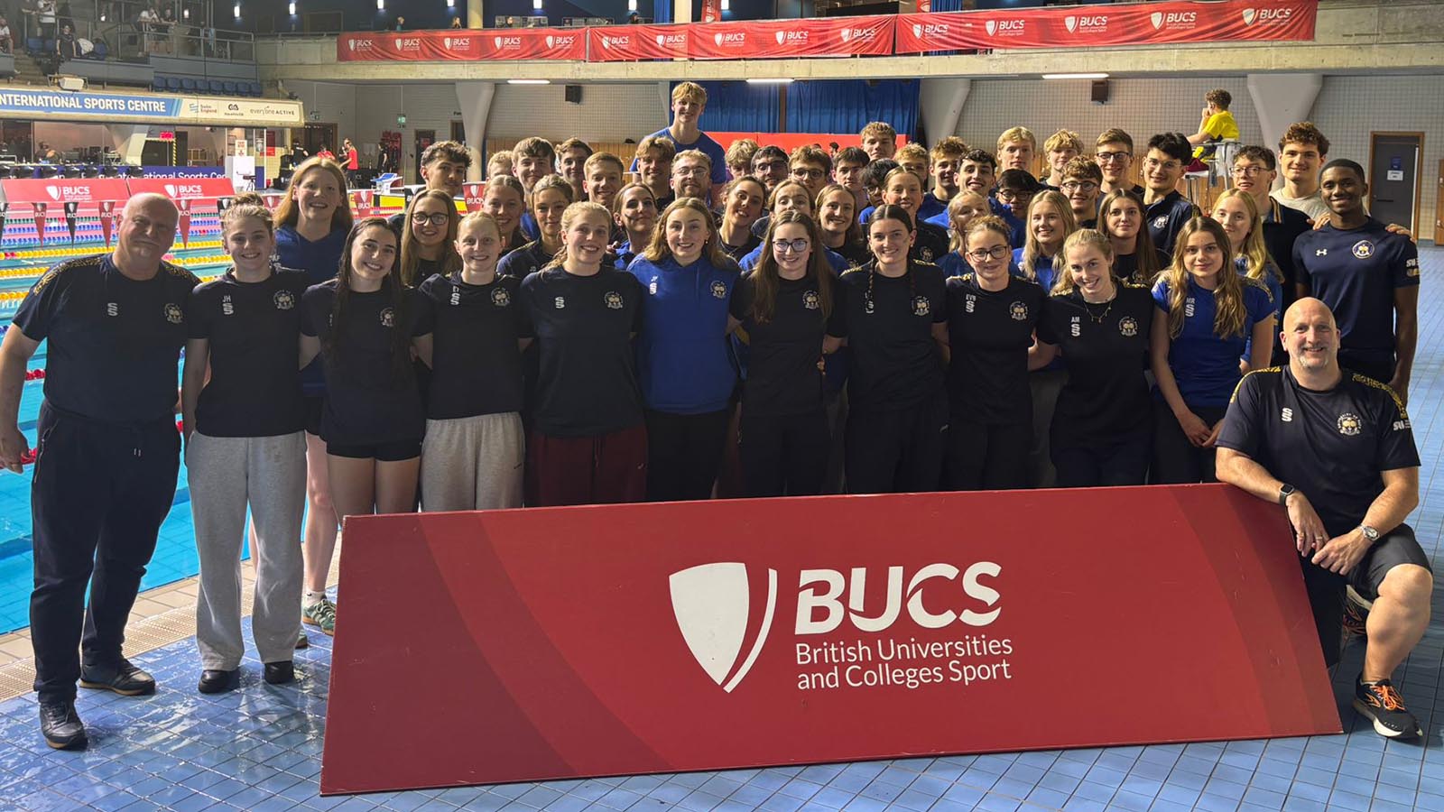 A squad picture of the University of Bath student swimming squad and coaches, standing behind red BUCS banner and underneath a scoreboard at Ponds Forge International Swimming Centre