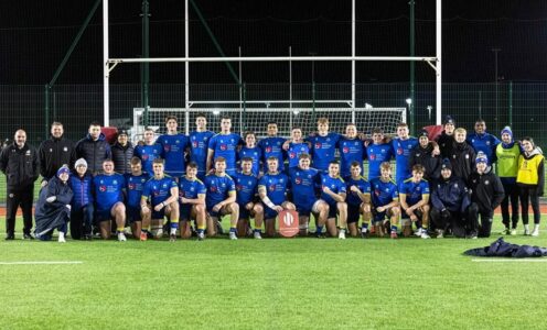 A squad photo of University of Bath men's 1st XV players, coaches and support staff in front of rugby posts. In front of captain Max Pearce, in the centre of the front row, is the BUCS Super Rugby Challenge Cup trophy.