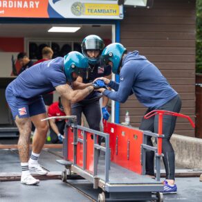 Three British Bobsleigh athletes enjoy a motivational huddle over a training cart underneath a Team Bath logo 