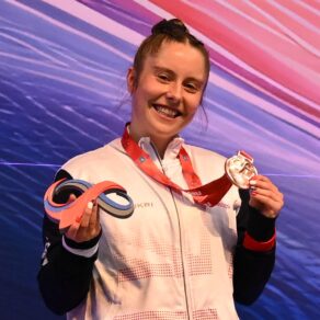 British Gymnastics' Kirsty Way has around her neck a bronze medal which she is lifting with her left hand. She has a trophy in her right hand. 