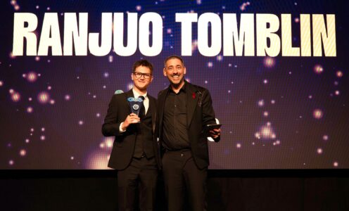 Artistic swimmer Ranjuo Tomblin and host Colin Murray on stage at the 2025 Aquatics GB Awards. They are both wearing suits and Ranjuo is holding a trophy. Behind them is an electronic display screen showing stars and Ranjuo Tomblin's name in bold letters.