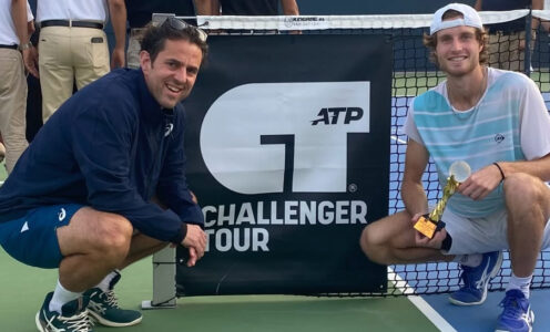 Team Bath Tennis player Toby Samuel, right, and his coach Ian MacDonald crouching down next to a tennis net featuring the ATP Challenger Tour logo. Toby is holding a trophy.