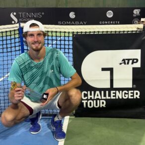Team Bath Tennis player Toby Samuel crouching down next to a tennis net featuring the ATP Challenger Tour logo. He is holding a trophy.