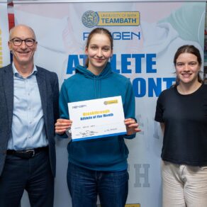 Emily Cox, holding a certificate, standing of front of an Athlete of the Month board. Standing either side of her are Stephen Baddeley, University of Bath Director of Sport, and Sarah Collin, Performance Sport Officer. 