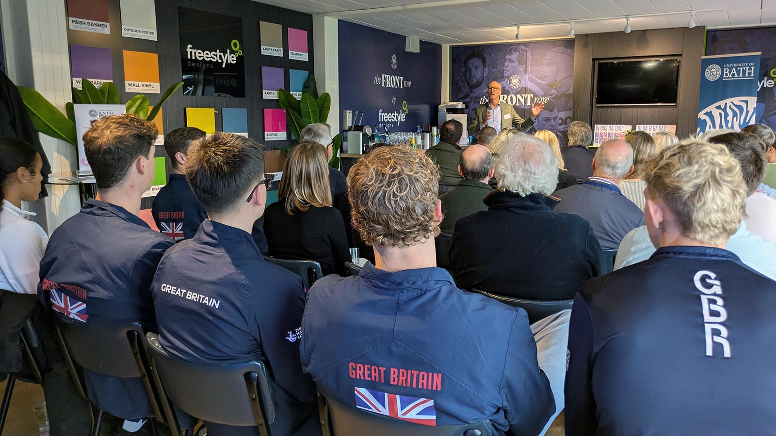 Stephen Baddeley speaks to an audience of people, whose backs we can see. In the foreground are four athletes wearing training tops with Great Britain or GBR on them.