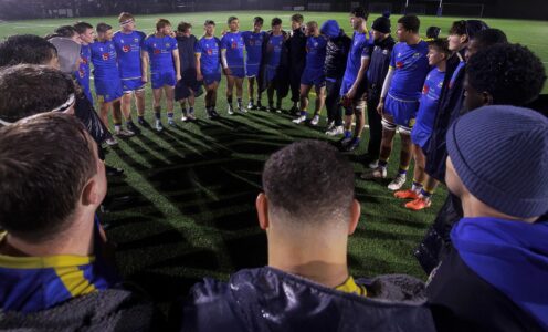 A group of rugby players standing in a circle, listening to a speech from their captain