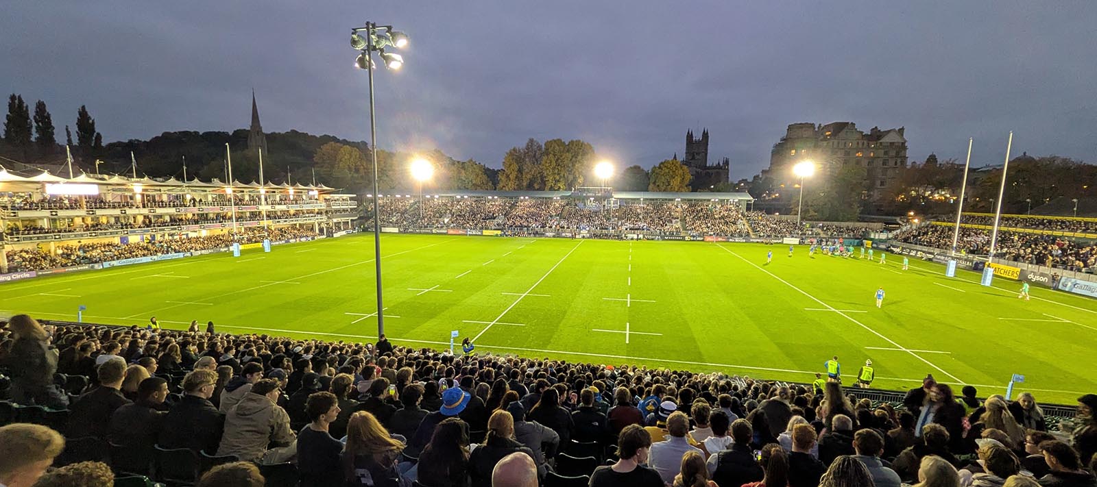 A panoramic view of The Rec rugby ground under floodlights, taken from the East Stand