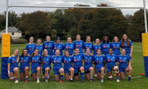 A squad picture of the University of Bath women's 1st XV, wearing blue kit, underneath rugby posts