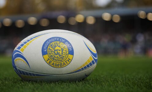 A close up of a rugby ball, branded with the Team Bath logo, on a pitch