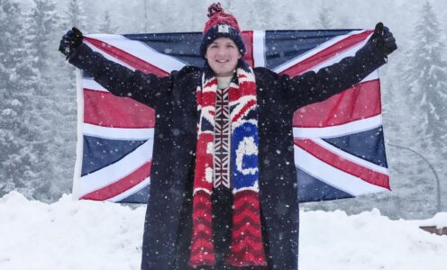 Bobsleigh pilot Brad Hall standing in snow, wearing Team GB kit and hat, holding a British flag behind his back with his arms outstretched.