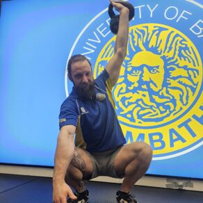 Matt Smith, a personal trainer at Team Bath, demonstrating a squat while lifting a weight with his left hand. Behind him is an illuminated Team Bath logo.