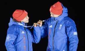 Tabby Stoecker and Matt Weston, both wearing blue Team GB coats and red woolly hats, clink gold medals on the podium
