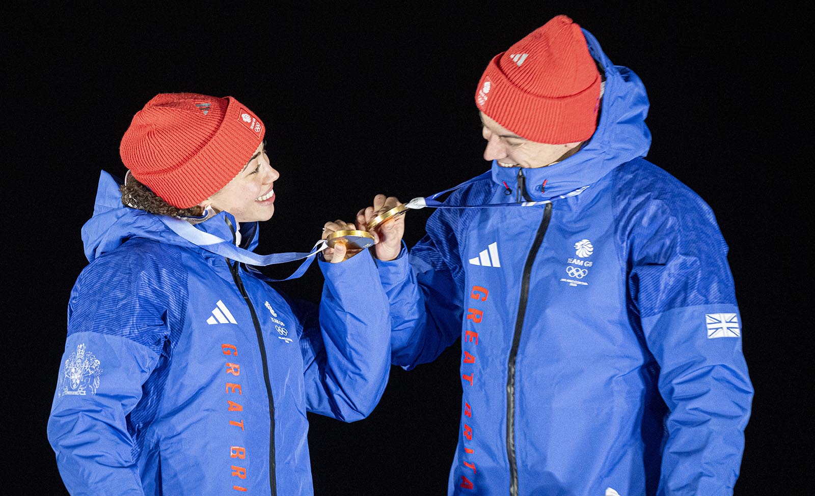 Tabby Stoecker and Matt Weston, both wearing blue Team GB coats and red woolly hats, clink gold medals on the podium