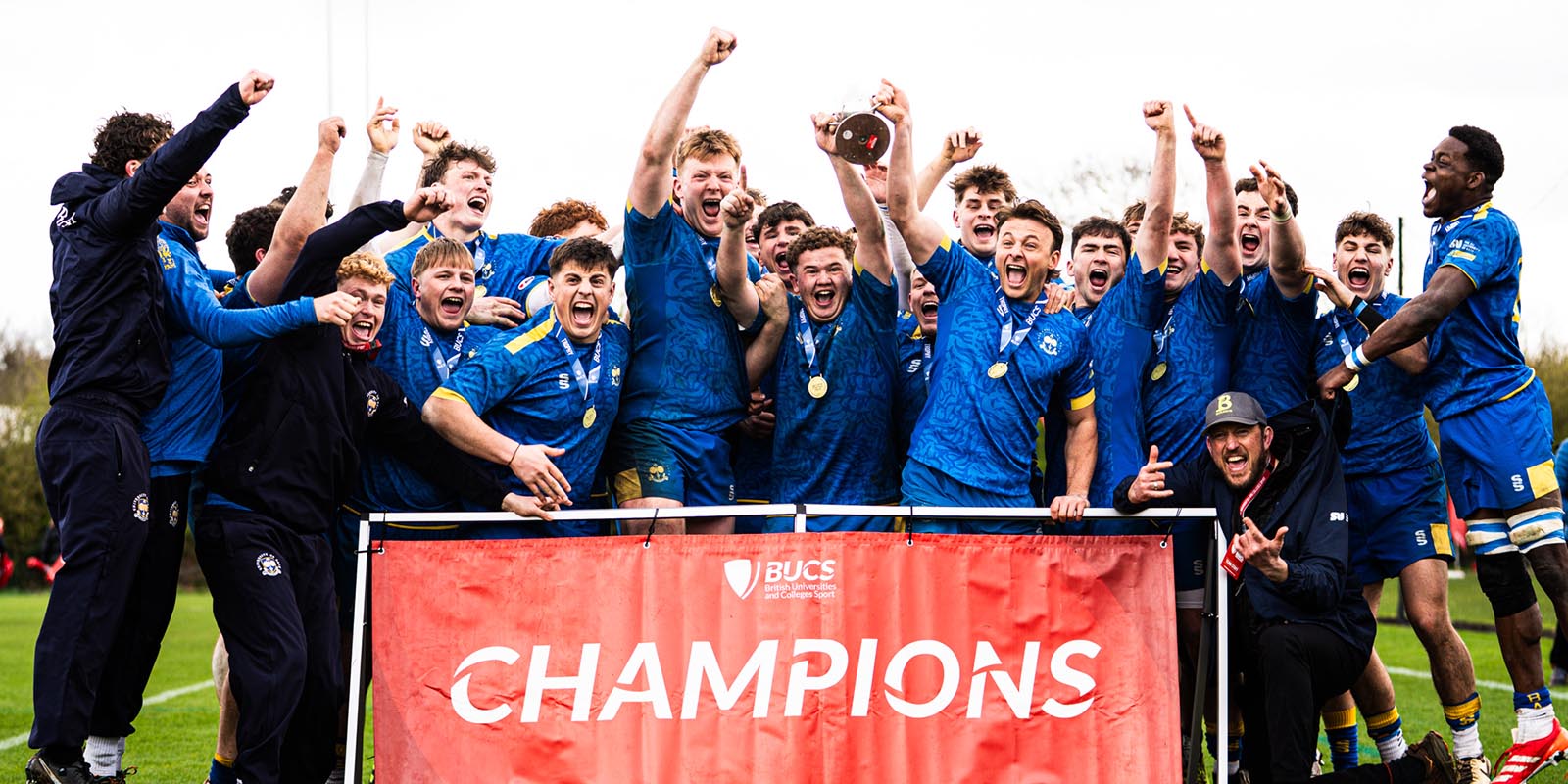The University of Bath men's 3rd XV rugby team and coaches celebrating behind a banner saying "Champions" as their captain (centre) holds aloft the BUCS National Trophy cup.
