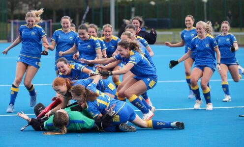 Joyous University of Bath women's hockey players rush to jump on top of their goalkeeper, lying on a blue pitch, after her save sealed victory in the BUCS National Vase final 2026