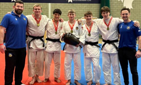 Coaches Adam Hall (left) and Gregg Varey with five Team Bath judoka who won the men's team title at the BUCS Judo Championships 2026. They are holding a black trophy.