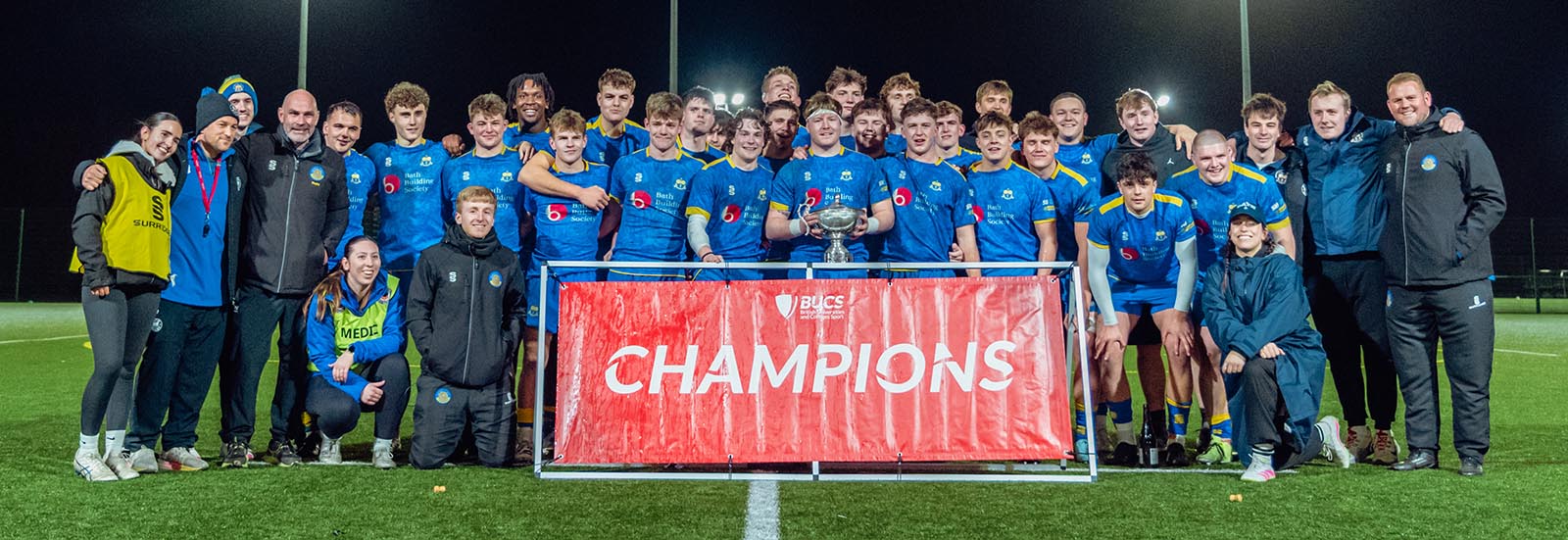 A squad shot of University of Bath Rugby Club players, coaches and support staff standing behind a Champions banner after winning the BUCS Super Rugby league title