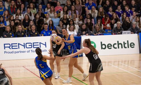 Team Bath Netball's Lottie Robinson shapes to pass the ball to team-mate Ellie Ervine as she is closed down by Loughborough Lightning defenders. Behind them is a stand full of spectators.