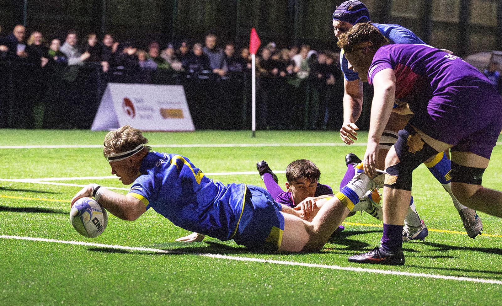 Max Pearce reaches his left arm out to touch down as he is tackled by a Loughborough player wearing purple.
