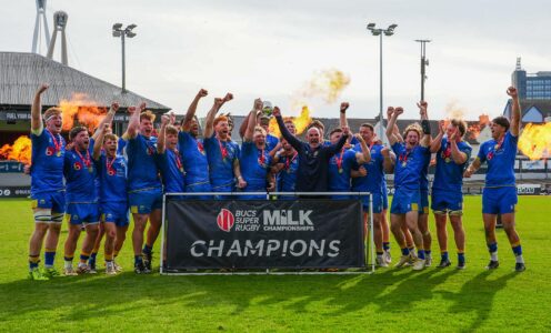 Captain Max Pearce and Head of Rugby Aaron James lift the BUCS Super Rugby Milk Championship trophy as their team-mates celebrate and fireworks go off behind them