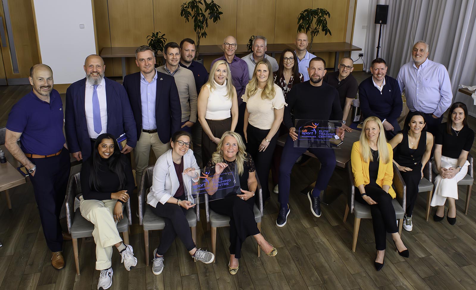 A group of people from UK Sport-accredited Elite Training Centres, sitting down and standing, looking at the camera