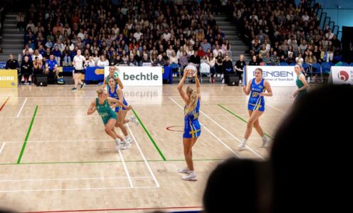 Team Bath Netball and LexisNexis Dragons players on court at the Team Bath Arena, as seen from the stands. Behind the players is another full stand of spectators.