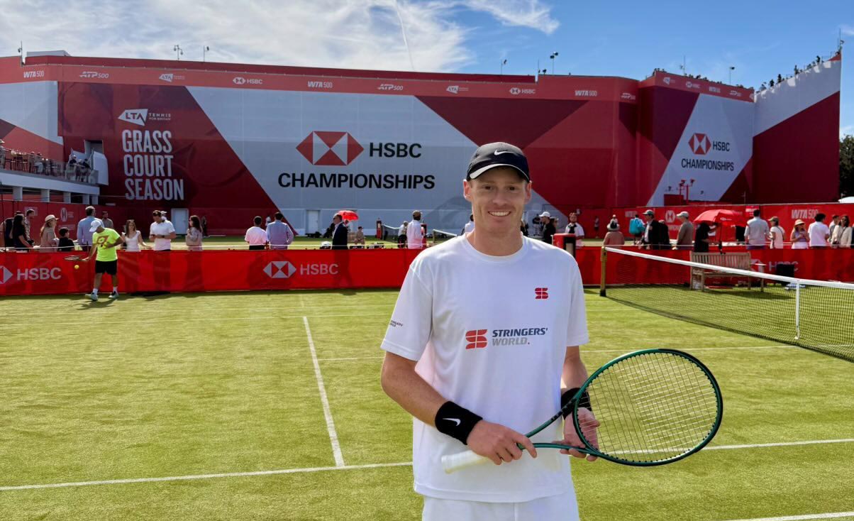 Tennis player Ben Jones standing on court, smiling at the camera, with the Queen's Club main stand behind him featuring the words 'HSBC Championships'.