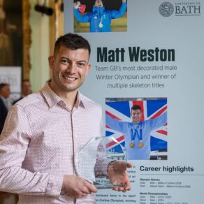 Matt Weston, holding a glass trophy, standing in front of a Hall of Fame panel featuring his name, photo and career achievements
