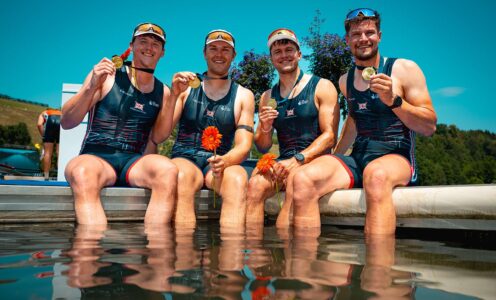 The four members of the GB Rowing Team men's quad, sitting with their feet in water while holding gold medals.