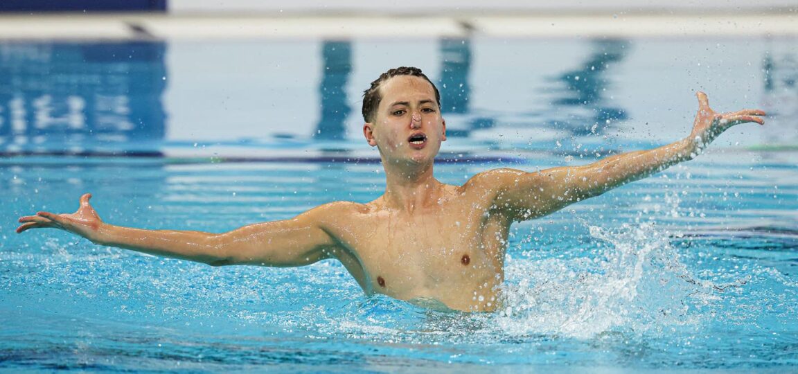 Artistic swimmer Ranjuo Tombin in the swimming pool during a free routine, his arms are outstretched.