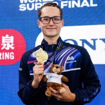 Artistic swimmer Ranjuo Tomblin, on the World Cup Super Final podium, holding a gold medal in his right hand and a mascot teddy bear in his left