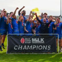 University of Bath Rugby Club players celebrate behind a banner saying "CHAMPIONS" as captain Max Pearce and head coach Aaron James lift the BUCS Milk Championship trophy