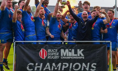 University of Bath Rugby Club players celebrate behind a banner saying "CHAMPIONS" as captain Max Pearce and head coach Aaron James lift the BUCS Milk Championship trophy