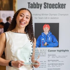 Tabby Stoecker, holding a glass trophy, standing in front of a Hall of Fame panel featuring her name, photo and career achievements