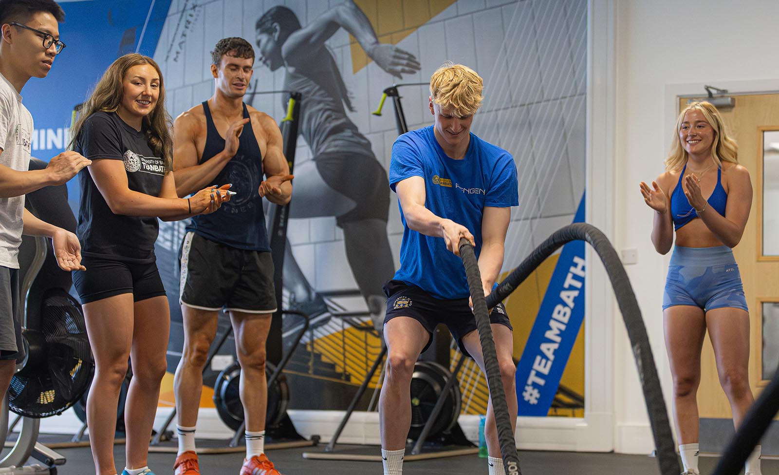 A gym user uses the battle ropes as colleagues give him encouragement