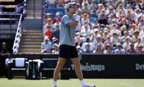 Team Bath Tennis player Toby Samuel on a grass court, clenching his fist in celebration, as a stand of spectators watches on behind him