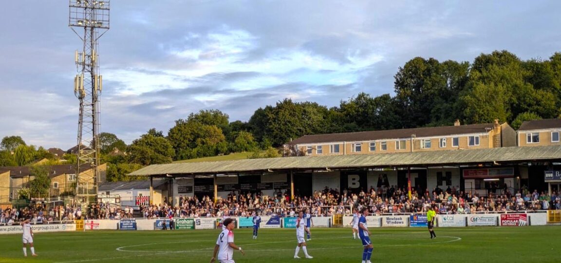 Two football teams playing on a pitch with a stand of spectators and old-style floodlights in the background.