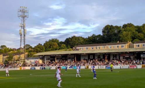 Two football teams playing on a pitch with a stand of spectators and old-style floodlights in the background.